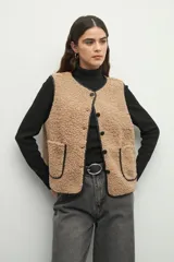 Chaleco color beige de corderito con cuello redondo, cierre frontal con botones a presión y dos bolsillos delanteros con ribete de cuero sintético negro.