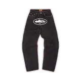 Pantalón de jeans negro, corte baggy, con pespuntes en color naranja. Presenta un estampado ovalado blanco en la parte frontal superior con un diseño de paisaje. Incluye etiqueta de marca en la cintura.