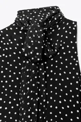 Vestido midi negro con estampado de lunares blancos, cuello halter con pañuelo integrado, sin mangas y bajo asimétrico. Cierre con abertura y botón en la nuca.