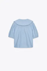 Camisa celeste a rayas verticales blancas, de corte recto, con cuello bobo con bordados y escote pico con lazada. Tiene mangas cortas abullonadas con elástico y cierre frontal con botones.
