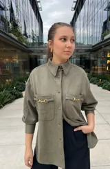 Camisa verde con cuello clásico, cierre frontal con botones, mangas 3/4 con puños abullonados, dos bolsillos con solapa en el pecho y detalle de cadena dorada en cuello y bolsillos.