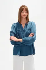 Blusa de lyocell en tono azul denim, con cuello en V, cierre frontal parcial con botones y mangas largas abullonadas con puños elásticos. Presenta un diseño holgado con detalle de fruncido en el pecho.