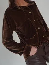 Camisa roja de terciopelo con cuello clásico, botones frontales y mangas largas con puños abotonados. Tiene dos bolsillos de parche en el pecho.
