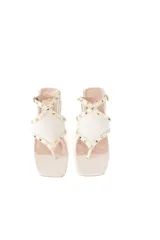 Sandalias de cuero color crema con tira entre los dedos, correa ajustable en el tobillo, taco bajo cuadrado y pequeñas tachas doradas.