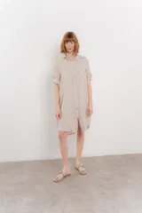 Vestido camisero corto color beige de lino 100%, con botones en la parte delantera, cuello camisero, mangas cortas con botón para arremangar y bolsillos laterales.