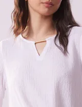 Blusa blanca de tejido texturado con cuello redondo, escote en V con aplique metálico y mangas 3/4 con puños elásticos. El ruedo se ata con un nudo.