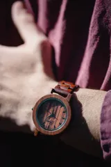 Reloj de pulsera con caja, esfera y malla de madera de sándalo rojo, con marcadores y manecillas negras. Presenta un diseño circular con corona lateral.