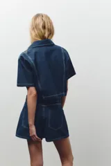 Camisa corta de denim azul con cuello solapa, escote pico, manga corta, bolsillos delanteros de plastrón y cierre frontal con botones metálicos.