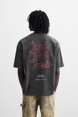 Camiseta negra con cuello redondo y mangas cortas. Estampado frontal con la frase "Cherry Pill Blush Delight" y una ilustración de una mujer en un cohete. Tiene un efecto de lavado que le da una apariencia única.
