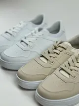 Zapatillas deportivas de simil cuero disponibles en colores blanco y beige, con diseño clásico.