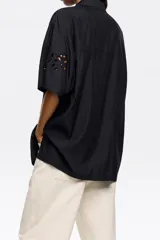 Camisa negra de manga corta con cuello clásico y cierre frontal con botones. Presenta bordado inglés con motivos florales. Tiene un bolsillo de parche en el pecho.