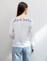 Blusa blanca de rayón con escote en V y mangas largas. Presenta bordados florales en tonos naranja, azul y negro en la parte delantera y en las mangas.