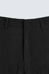 Pantalón negro de corte relajado (relaxed fit) con textura arrugada, tiro medio, bolsillos laterales y bolsillos traseros de vivo. Cierre frontal con cremallera y gancho metálico.