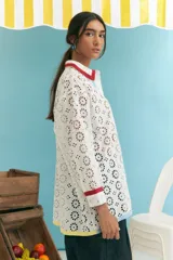 Camisa blanca oversize de algodón con bordado calado tipo broderie, cuello camisero con ribete rojo, cierre frontal con botones y mangas largas con puños con ribete rojo. Presenta una franja vertical con ribete celeste y otra con ribete amarillo.