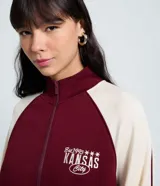Saco femenino de poliviscosa, color blanco con mangas raglán marrones y ribetes blancos. Tiene cuello alto, cierre frontal de cremallera y bordado lettering "Kansas City" en el pecho.