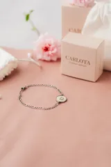 Pulsera de acero quirúrgico plateado con cadena de eslabones y dije circular blanco con borde negro y la letra "D" en negro.