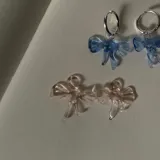 Par de aros colgantes con argolla de acero inoxidable y dije de vidrio con forma de moño, disponibles en color azul y rosa.