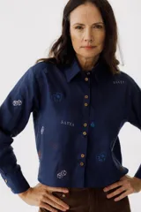Camisa azul de lino con cuello clásico, manga larga y cierre frontal con botones. Presenta bordados de dados, cartas y el nombre de la marca.