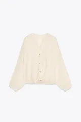 Blusa color crema de tejido arrugado con hilo metalizado, cuello redondo con escote en pico, manga larga abullonada y nudo delantero. Cierre frontal con botones con flores bordadas.