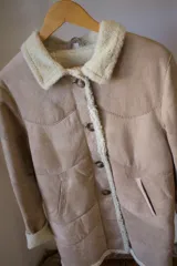 Abrigo gamulán marrón con cuello, solapa, puños y borde frontal de piel sintética blanca. Tiene cierre con botones y bolsillos laterales.