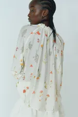 Blusa oversized de viscosa color crema con estampado floral. Tiene cuello corto con volado y abertura en pico con tiras finas. Las mangas son raglán largas con elástico fino en los puños y el bajo es redondeado.