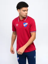 Remera tipo polo roja con cierre corto, cuello y puños con detalles tricolor. Incluye logo de Umbro y escudo del Club Nacional de Football. Confeccionada en tejido de punto.