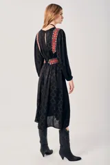 Vestido largo negro con estampado de hojas en relieve, mangas largas abullonadas y falda plisada. Presenta detalles bordados en rojo, blanco y azul en los hombros y la cintura.