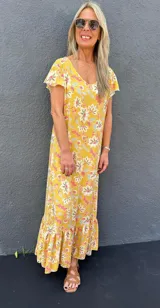 Vestido largo amarillo con estampado floral en tonos rosa, blanco y celeste. Tiene mangas cortas con volados y un volante en el ruedo.