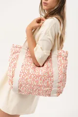 Bolso tipo tote bag de tela color crudo con estampado de letras en color naranja. Tiene asas de hombro de tela con diseño de espiga y un bolsillo frontal.