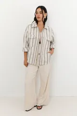 Chaqueta de algodón a rayas verticales en tonos beige y gris, con cuello camisero, cierre frontal con botones y mangas largas con puños abotonados. Tiene bolsillos delanteros con solapa y galones ondeados a tono.