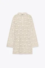Vestido corto color crudo confeccionado en tejido tipo crochet con diseño geométrico floral. Tiene cuello solapa, abertura con botón en la parte superior y mangas largas acampanadas.