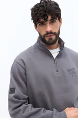 Buzo de algodón gris oscuro con cuello alto y cierre frontal parcial. Presenta un parche de marca en la manga izquierda y un logo bordado en el pecho.