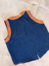 Chaleco de jean azul con detalles de cuero color suela en cuello y sisas, con botón de madera en la parte superior trasera.