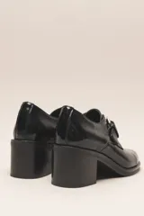 Zapato de cuero vacuno charol negro, con punta redondeada y detalle de perforado en la capellada. Tiene una tira con hebilla metálica y un taco cuadrado de 4 cm.