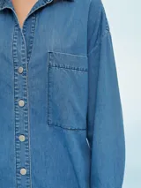 Camisa de denim negro con corte oversize, cuello clásico, cierre frontal con botones metálicos y bolsillo de parche en el pecho.