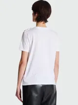 Remera blanca de algodón con cuello redondo y mangas cortas. Estampado frontal con el logo 'BALMAIN PARIS' en color negro. Detalle de botones dorados en el hombro izquierdo.