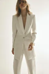 Blazer larga color blanco, de cuello solapa y manga larga con hombreras. Tiene bolsillos delanteros, forro interior y cierre frontal con botón metálico en relieve.