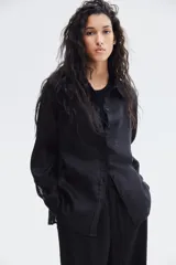 Camisa negra de lino liviano, con cuello clásico, cierre frontal con botones, canesú de doble capa en la espalda, hombros ligeramente caídos, mangas largas con puños abotonados, bolsillo de parche en el pecho y bajo redondeado.