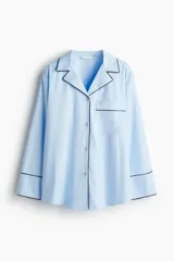 Camisa oversized de algodón color celeste claro con ribetes en contraste negro. Presenta cuello abierto, cierre frontal de botones, un bolsillo superior de parche y mangas largas con puños anchos.