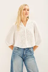 Camisa blanca de manga larga con cuello redondo y bordado floral en el delantero. El delantero es de algodón plano, mientras que las mangas y la espalda son de jersey a tono. Presenta un efecto fruncido en el centro de la espalda y puños en las mangas largas.