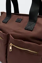 Bolso tote marrón de nylon con doble asa de mano negra y correa bandolera negra regulable y desmontable. Tiene dos bolsillos frontales con cierre metálico dorado.