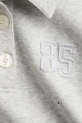 Remera gris con cuello polo, de corte cropped y manga larga. Presenta apliques de strass y bordado con el número 85 en el pecho.