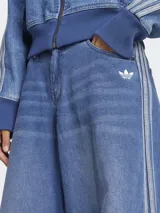 Pantalón de jean Adidas de corte ancho, color azul denim con tres tiras verticales plateadas a los costados y logo bordado en blanco en el bolsillo delantero.