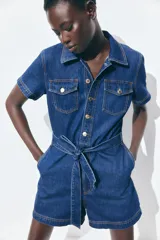 Mono corto de denim azul, con cuello camisero, cierre frontal con botones, mangas cortas, bolsillos superiores de parche con botón, bolsillos al bies y bolsillos insertados atrás. Incluye cinturón extraíble para atar en la cintura.