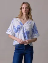 Blusa blanca estampada con flores azules, escote en V y mangas cortas amplias. Detalles de puntilla en escote y mangas.