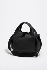Bolso tipo hobo o tote de nylon negro, con asas de mano cortas y una correa bandolera ajustable y desmontable de tejido negro. Presenta bolsillos laterales con cierre de cremallera y el logo de Bimba y Lola en blanco en la parte frontal inferior.