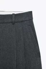 Pantalón negro de tiro alto con trabillas, bolsillos laterales y pinzas. Cierre frontal con cremallera, botón interior y gancho metálico.