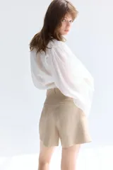 Short color beige de sarga con pinzas, tiro alto, cinturón trenzado ancho con hebilla circular, cierre con botón y corchete ocultos, bolsillos laterales discretos y bolsillos traseros insertados.