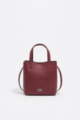 Bolso pequeño de piel color borgoña, con estructura rígida y forma de cubo. Posee doble asa de mano y una correa larga para llevar al hombro o cruzado. El frente lleva el logo metálico de Bimba y Lola.
