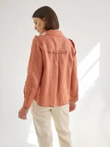 Camisa blanca de lino con cuello clásico, abotonada al frente con botones de madera, mangas largas con volados en los hombros y corte femenino.